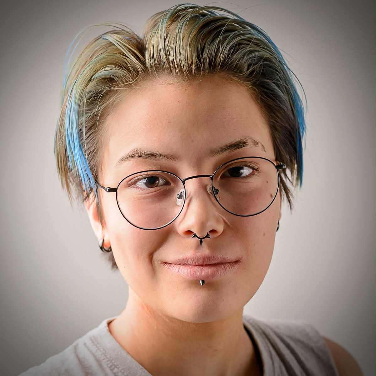 close up picture taken from the front.
                    The person on the camera is wearing glasses and have a few piercings as well as dyed blue hair.
                    their eyes are brown and they are wearing a grey shirt